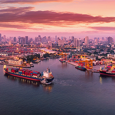 International port with large shipping container sailing away down the river, background of a large city and foreground showing equipment, containers and buildings of the port