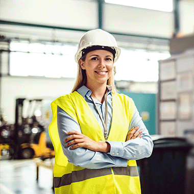 Woman in a white hard hat and yellow high viz vest with crossed arms smiling and looking determined, warehouse in the background